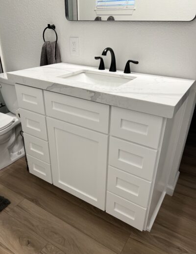 A white bathroom vanity with a marble countertop and single basin sink, featuring several drawers and a cabinet. The sink has a black faucet. A hand towel hangs on a nearby towel ring.