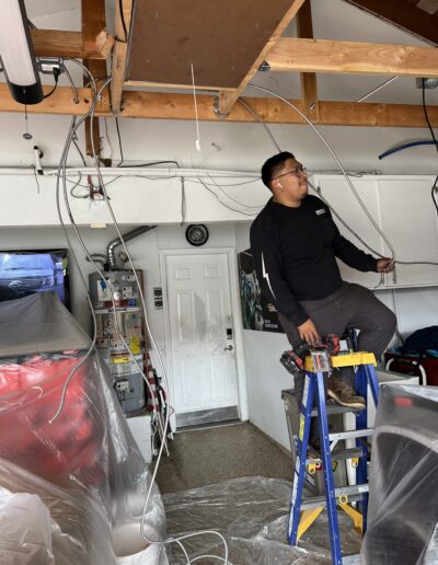 A person stands on a blue ladder working with wires in a garage. Various tools, plastic-covered items, and a furnace are visible in the background.