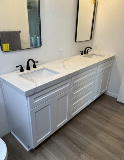 A bathroom features a double-sink vanity with white cabinetry and marble countertop. Two rectangular mirrors and black faucets are mounted above the sinks. Light wood flooring is visible.