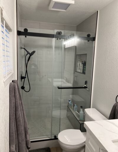 A modern bathroom featuring a glass-enclosed shower with a black showerhead, gray towels on a hook, a white countertop sink, and various toiletries on a glass shelf inside the shower.