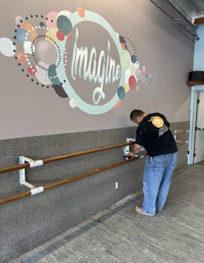 A man in a black t-shirt installs a wooden handrail below a mural with the word "Imagine" on a wall inside a room.