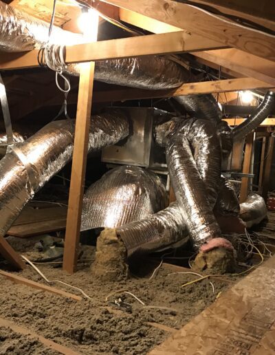 An attic with exposed wooden beams, ductwork covered in shiny insulation, and bare insulation material spread on the floor. Light fixtures are attached to the wooden beams overhead.