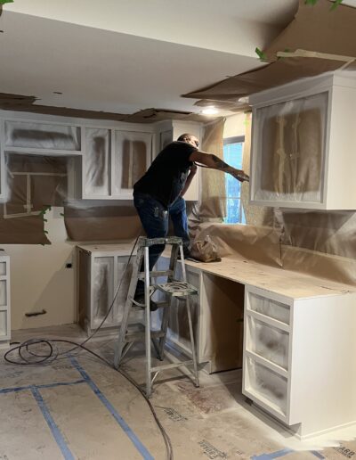 A person on a stepladder spray paints kitchen cabinets while the room is covered in protective paper and tape.