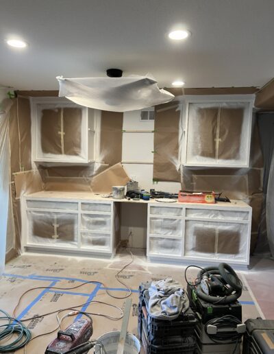 A work-in-progress kitchen renovation with white cabinets and drawers partially covered in protective plastic, tools and equipment scattered on the countertop and floor, and construction materials visible around the area.