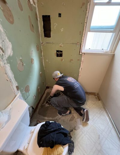 A person wearing a backwards cap kneels on the floor of a bathroom under renovation, working on plumbing or tiling near an exposed wall. A toilet and cleaning supplies are nearby.