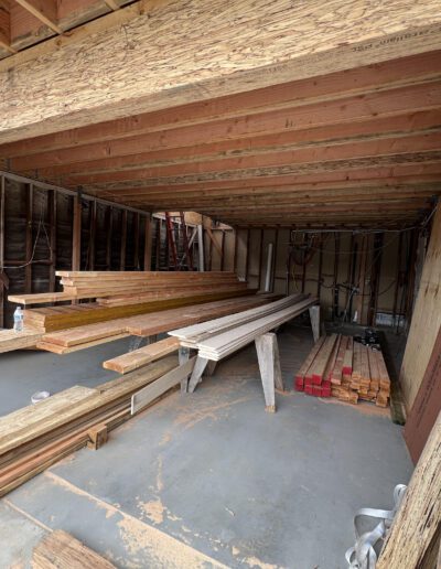 A construction site featuring stacks of wooden planks and boards stored under a partially covered structure with exposed beams. Tools and other materials are scattered on the concrete floor.