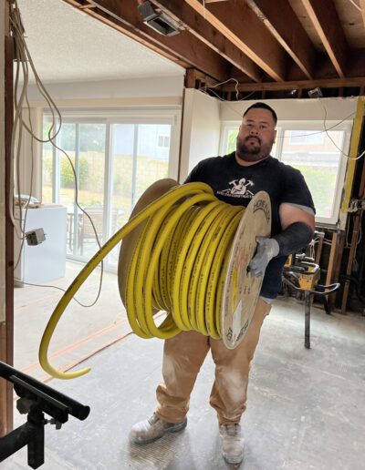 A construction worker carrying a large spool of yellow cable in a partially renovated interior space with exposed beams and wiring.