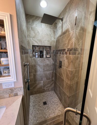 Modern shower with beige tiled walls, mosaic accent, built-in shelf with toiletries, overhead rain showerhead, handheld showerhead, and a grab bar. Adjacent sink with shelving unit visible.