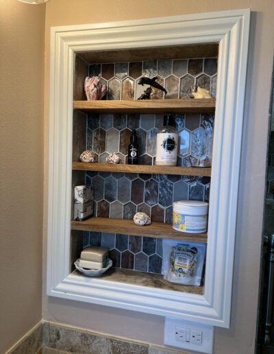 A recessed shelf with honeycomb-patterned tiles contains various personal care items including soap, lotions, and decorative objects. The shelf has a white frame and is built into a beige wall.