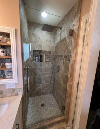 A modern shower with stone tile walls and flooring, a glass door, a square overhead showerhead, and built-in shelves containing toiletries.